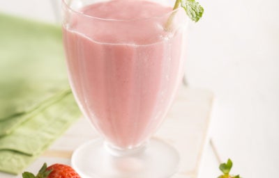 A imagem mostra um copo de smoothie rosa de morango, decorado com um raminho de hortelã fresca. O copo é transparente e tem uma base fosca, colocado sobre uma superfície de madeira clara. Ao redor, há três morangos frescos