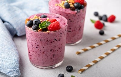 Fotografia em tons claros com dois copos de vidro ao centro. Dentro dos dois copos existe uma receita de smoothie de frutas vermelhas de cor rosa arroxeada. Por cima do smoothie, existe várias frutas vermelhas diversas.