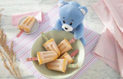 Foto da receita de Picolézinho de Manga com Banana. Observa-se um potinho verde claro com 5 picolés laranjas dentro, com cabinhos de madeira decorados. O pote está sobre um paninho listrado colorido. Foto da receita de Picolézinho de Manga com Banana. Observa-se um potinho verde claro com 5 picolés laranjas dentro, com cabinhos de madeira decorados. O pote está sobre um paninho listrado colorido.
