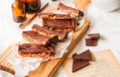 Fotografia em tons de marrom em uma bancada de madeira clara, uma tábua de madeira, barras de chocolate com pasta de amendoim ao centro e pedaços de chocolate soltos ao lado.
