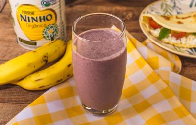 Fotografia em tons de amarelo de uma bancada com um pano xadrez amarelo e branco e um copo com a bebida de açaí. Ao fundo bananas, uma lata de NINHO e um prato com uma crepioca.