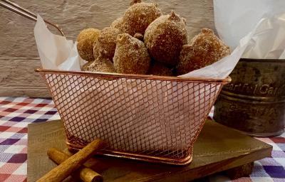 Foto da receita de Bolinho de Chuva. Observa-se uma cesta de metal com guardanapo e os bolinhos de chuva dentro sobre uma tábua de madeira em cima de uma toalha quadriculada.