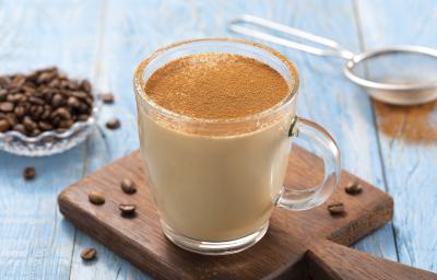 Foto da receita de Cappuccino Quente Cremoso. Observa-se uma caneca de vidro transparente com o cappuccino polvilhado de canela em cima de uma pequena tábua de madeira sobre uma superfície de madeira azul clara. Ao fundo, grãos de café e uma peneira.