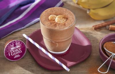 Foto da receita de iced coffee com banana e cacau servida em um copo largo de vidro sobre um prato vinho com um canudo listrado, em cima de uma mesa de madeira com uma banana e um paninho roxo ao fundo