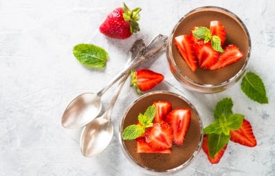 Fotografia em tons claros e vista de ciam de dois copos decorativos de tamanho normal com a sobremesa de taça moranguda, finalizada com chocolate e decoração de morangos em pedaços e folhas de hortelã.