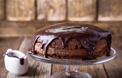 Fotografia de um bolo de chocolate em um suporte de vidro, coberto com ganache escura que escorre pelas laterais. Ao lado, um pequeno jarro de cerâmica com ganache, em um cenário acolhedor de madeira.