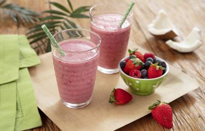 Fotografia em tons de vermelho, em uma bancada de madeira de cor marrom. Ao centro, uma bandeja de madeira com dois copos em cima contendo os smoothies. Ao lado, há um pano verde, uma tigela com morango e ao fundo algumas folhas e um coco partido ao meio.