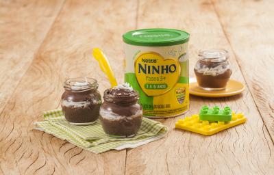 Fotografia em tons de verde e amarelo em uma bancada de madeira clara, um paninho verde xadrez com dois potinhos de acrílico com a sobremesa de prestígio dentro dele. Ao lado, brinquedinhos, uma lata de Ninho Fases e mais um potinho com a sobremesa. Fotografia em tons de verde e amarelo em uma bancada de madeira clara, um paninho verde xadrez com dois potinhos de acrílico com a sobremesa de prestígio dentro dele. Ao lado, brinquedinhos, uma lata de Ninho Fases e mais um potinho com a sobremesa.