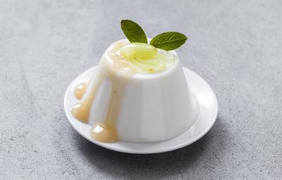 Fotografia de uma mousse de uva verde, com um creme e uma unidade de uva em cima, e para decorar, duas folhas de hortelã. A sobremesa está em um prato pequeno de sobremesa na cor branco, que está sobre uma mesa cinza.