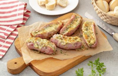 Foto em tons de bege e cor de rosa da receita de linguiça recheada servida em 5 porções sobre uma tábua de madeira com uma faca prateada ao lado. Ao fundo há um prato branco com fatias de pão francês e um pano listrado Foto em tons de bege e cor de rosa da receita de linguiça recheada servida em 5 porções sobre uma tábua de madeira com uma faca prateada ao lado. Ao fundo há um prato branco com fatias de pão francês e um pano listrado