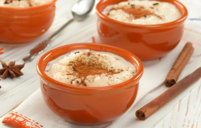 Fotografia em tons claros de três recipientes fundos e pequenos marrons sobre uma mesa clara com alguns paus de canela e cravos da índia. Dentro dos recipientes têm arroz doce com canela.