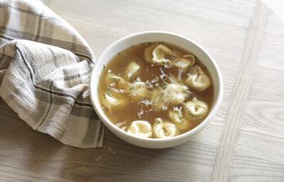 Fotografia de um recipiente branco fundo com uma sopa de capeletti, sobre uma mesa de madeira. Ao lado, um pano branco com linhas na cor cinza e preto. Fotografia de um recipiente branco fundo com uma sopa de capeletti, sobre uma mesa de madeira. Ao lado, um pano branco com linhas na cor cinza e preto.