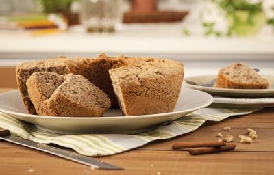 Fotografia em tons de marrom em uma mesa de madeira com um prato raso branco apoiado em um pano cinza listrado em bege e o bolo de maçã e nozes. Ao lado, um prato com um pedaço do bolo e paus de canela espalhadas pela mesa. Fotografia em tons de marrom em uma mesa de madeira com um prato raso branco apoiado em um pano cinza listrado em bege e o bolo de maçã e nozes. Ao lado, um prato com um pedaço do bolo e paus de canela espalhadas pela mesa.