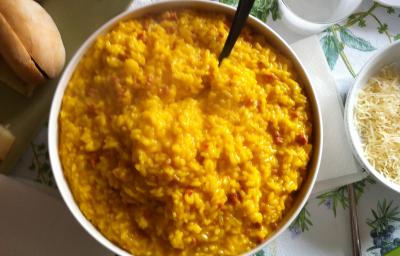 Fotografia vista de cima de um refratário fundo branco com arroz cremoso com açafrão. Ao lado direito, dois pratos e dois copos brancos, um bowl pequeno branco com queijo ralado, e no outro lado, um prato quadrado verde com alguns pães. Fotografia vista de cima de um refratário fundo branco com arroz cremoso com açafrão. Ao lado direito, dois pratos e dois copos brancos, um bowl pequeno branco com queijo ralado, e no outro lado, um prato quadrado verde com alguns pães.