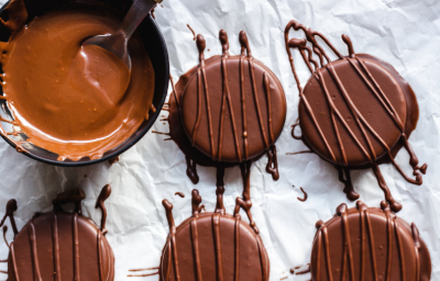 Imagem mostra 5 alfajores redondos cobertos por chocolate ao leite escuro, dispostos sobre um papel manteiga branco. Ao lado um recipiente com chocolate derretido e uma colher.