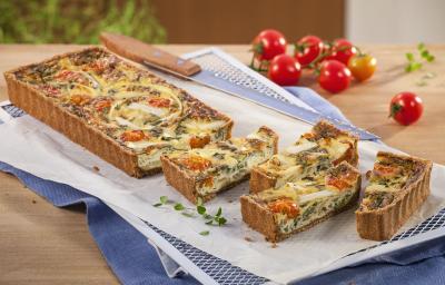 Fotografia em tons branco, dourado e vermelho, a direita e ao alto tomates-cereja e folhas de tomilho, ao centro travessa branca com quiche assada e fatiada, sobre papel manteiga e guardanapo azul em bancada de madeira. Fotografia em tons branco, dourado e vermelho, a direita e ao alto tomates-cereja e folhas de tomilho, ao centro travessa branca com quiche assada e fatiada, sobre papel manteiga e guardanapo azul em bancada de madeira.
