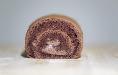 Fotografia de um bolo de rolo de chocolate, em tons de marrom, ao centro de uma mesa de madeira clara. Fotografia de um bolo de rolo de chocolate, em tons de marrom, ao centro de uma mesa de madeira clara.