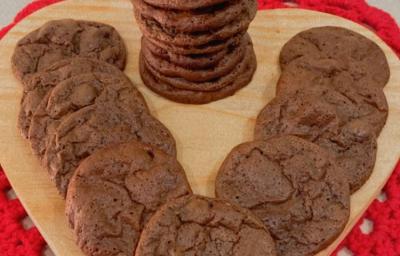 Fotografia de uma tábua de madeira em formato de coração sobre um pano vermelho. Sobre a tábua tem uma torre de uma receita de cookie brownie, ao lado de mais unidades do cookie completando o espaço da tábua. Fotografia de uma tábua de madeira em formato de coração sobre um pano vermelho. Sobre a tábua tem uma torre de uma receita de cookie brownie, ao lado de mais unidades do cookie completando o espaço da tábua.