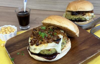 Fotografia de um hambúrguer completo com pão, cebola caramelizada, hambúrguer vegetal de cor esverdeada e queijo derretido. Ao fundo existe outro hambúrguer igual e ao lado um pequeno copo com molho de cor escura.
