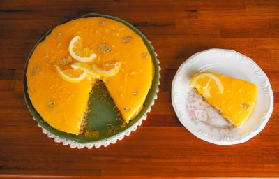 Fotografia mostra cheesecake de limão siciliano visto de cima, com cobertura amarelo vibrante e fatias de limão. Uma fatia servida ao lado em prato branco decorado.