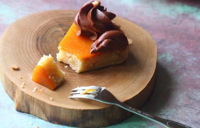 Fotografia de uma fatia de torta com damasco e mousse de chocolate por cima decorado por um bico de confeitar sobre uma tábua redonda de madeira e um garfo apoiado sobre. Ao fundo, a torta inteira sobre outra tábua de madeira redonda. Fotografia de uma fatia de torta com damasco e mousse de chocolate por cima decorado por um bico de confeitar sobre uma tábua redonda de madeira e um garfo apoiado sobre. Ao fundo, a torta inteira sobre outra tábua de madeira redonda.