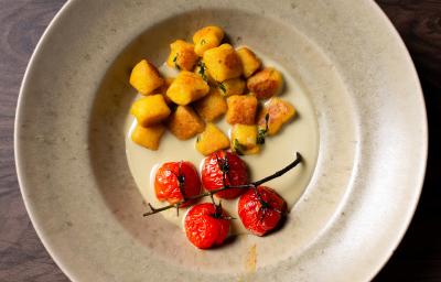 Fotografia em tons de branco com um prato branco ao centro. Dentro do prato existe uma receita de nhoque de abóbora acompanhada de tomates confitados