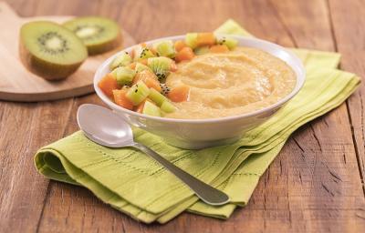 Foto em tons de verde da receita de creminho de laranja servida em um bowl de porcelana branco com uma colher prateada ao lado, sobre um pano verde claro. Ao fundo, há uma tábua de madeira com kiwis cortados em cima Foto em tons de verde da receita de creminho de laranja servida em um bowl de porcelana branco com uma colher prateada ao lado, sobre um pano verde claro. Ao fundo, há uma tábua de madeira com kiwis cortados em cima