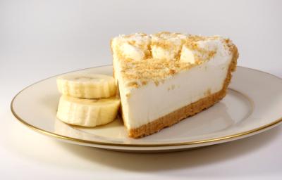 Fotografia de uma fatia de torta de banana e leite MOÇA, com uma farofinha de biscoito e rodelas de banana por cima. A torta está em um prato de vidro branco com as bordas na cor dourado, com duas rodelas de banana ao lado da torta, sobre uma mesa branca. Fotografia de uma fatia de torta de banana e leite MOÇA, com uma farofinha de biscoito e rodelas de banana por cima. A torta está em um prato de vidro branco com as bordas na cor dourado, com duas rodelas de banana ao lado da torta, sobre uma mesa branca.
