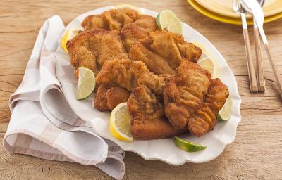Fotografia em tons de amarelo em uma bancada de madeira clara, um pano branco com listras bege, um prato branco oval com vários bifes suínos à milanesa. Ao fundo, pratos redondos amarelos com talheres em cima deles. Fotografia em tons de amarelo em uma bancada de madeira clara, um pano branco com listras bege, um prato branco oval com vários bifes suínos à milanesa. Ao fundo, pratos redondos amarelos com talheres em cima deles.