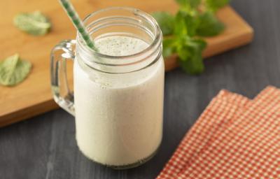 Fotografia em tons de branco, em uma bancada de madeira de cor preta. Ao centro, um copo com o suco de melão com um canudo verde dentro. Ao lado do copo, há um pano quadriculado e ao fundo, uma tábua de madeira de cor marrom folhas de hortelã espalhadas. Fotografia em tons de branco, em uma bancada de madeira de cor preta. Ao centro, um copo com o suco de melão com um canudo verde dentro. Ao lado do copo, há um pano quadriculado e ao fundo, uma tábua de madeira de cor marrom folhas de hortelã espalhadas.