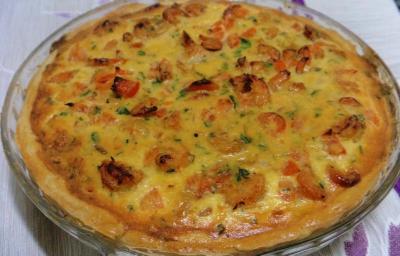 Foto em tons de amarelo da receita de quiche de camarão servida em uma porção grande em uma travessa circular de vidro sobre uma mesa com toalha branca e lilás Foto em tons de amarelo da receita de quiche de camarão servida em uma porção grande em uma travessa circular de vidro sobre uma mesa com toalha branca e lilás