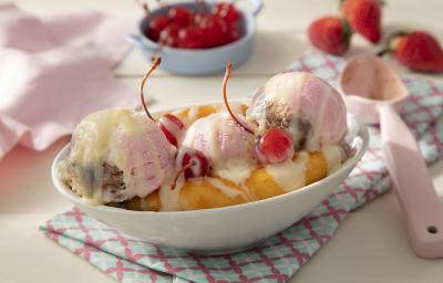 Fotografia em tons de rosa em uma bancada de madeira de cor marrom. Ao centro, uma toalha colorida e em cima, um pote contendo a banana split. Ao lado, uma colher rosa e ao fundo, um pano rosa, morangos e cerejas espalhados. Fotografia em tons de rosa em uma bancada de madeira de cor marrom. Ao centro, uma toalha colorida e em cima, um pote contendo a banana split. Ao lado, uma colher rosa e ao fundo, um pano rosa, morangos e cerejas espalhados.