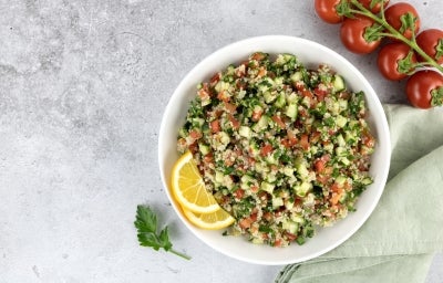 Fotografia em tons claros vista de cima de um recipiente fundo, branco, com uam receita de tabule. O recipiente está ao lado de alguns tomates, um pano verde claro e folhas de salsinha, sobre uma mesa de cimento queimado e tom claro.