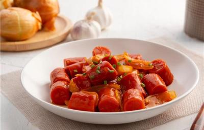 Fotografia em tons claros de um prato de vidro fundo com salsichas em pedaços e um pouco de molho com cebolas cortadas em tiras médias, cozidas no molho também. O prato está sobre um pano fino bege, o qual está apoiado em uma mesa branca. Fotografia em tons claros de um prato de vidro fundo com salsichas em pedaços e um pouco de molho com cebolas cortadas em tiras médias, cozidas no molho também. O prato está sobre um pano fino bege, o qual está apoiado em uma mesa branca.