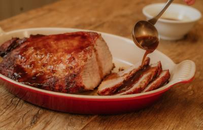 Fotografia em tons de marrom de uma bancada de madeira, sobre ela um recipiente refratário vermelho e branco, sobre ele um pernil com calda. Ao fundo um recipiente redondo e branco com calda.