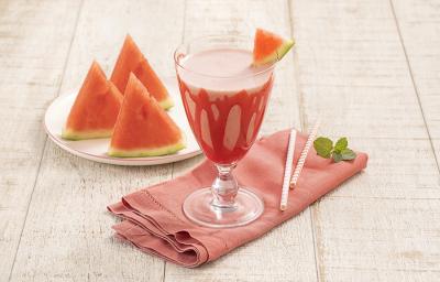 Foto da receita da bebida refrescante de melancia e morango servida em uma taça de vidro com um pedaço de melancia para decorar, há um pano vermelho embaixo da taça e sobre a mesa de madeira clara. Ao fundo um pires branco com três pedaços de melancia