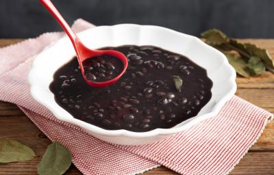 Fotografia em tons de preto e vermelho de uma bancada de madeira, um pano com listras vermelhas e brancas, uma travessa redonda com feijão cozido e seu caldo e uma concha vermelha com um pouco do feijão acima da travessa. Fotografia em tons de preto e vermelho de uma bancada de madeira, um pano com listras vermelhas e brancas, uma travessa redonda com feijão cozido e seu caldo e uma concha vermelha com um pouco do feijão acima da travessa.