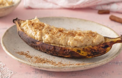 Imagem mostra uma banana assada em tons dourados e marrons, aberta com um creme e pedaços de amendoim