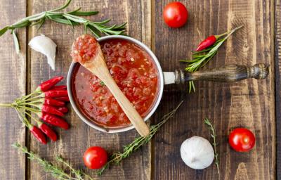 Fotografia vista de cima de um molho de tomate caseiro picante dentro de uma panela, e uma colher de madeira apoiada em cima. Ao redor, tomates inteiros, pimentas, ramos de alecrim e cebolas espalhadas sobre uma mesa de madeira. Fotografia vista de cima de um molho de tomate caseiro picante dentro de uma panela, e uma colher de madeira apoiada em cima. Ao redor, tomates inteiros, pimentas, ramos de alecrim e cebolas espalhadas sobre uma mesa de madeira.