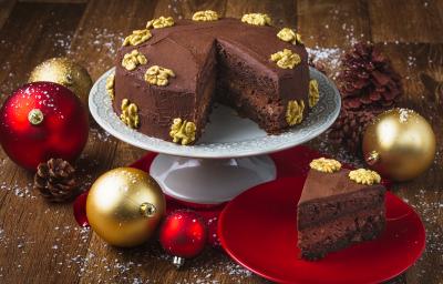 Foto da receita de Bolo Cremoso de Chocolate e Nozes, em um prato branco alto e uma fatia em um prato vermelho. Tudo está em uma bancada de madeira escura decorada com bolas de natal, pinhas e neve artificial Foto da receita de Bolo Cremoso de Chocolate e Nozes, em um prato branco alto e uma fatia em um prato vermelho. Tudo está em uma bancada de madeira escura decorada com bolas de natal, pinhas e neve artificial