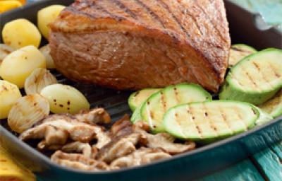 Fotografia em tons de azul e amarelo em uma bancada de madeira azul, um pano amarelo, uma frigideira com a picanha, abobrinha, alho e cebola dentro dela.
