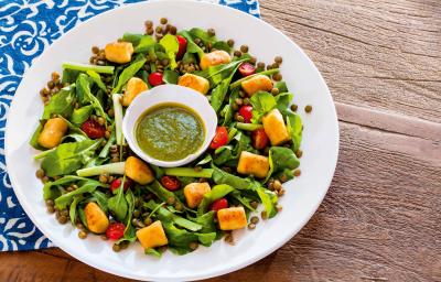 Fotografia em tons de verde em uma bancada de madeira com um pano azul desenhado, um prato branco raso redondo com a salada de cenoura, rúcula e tomate cereja.