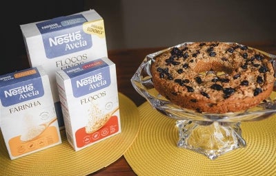 Fotografia em tons claros de três embalagens de aveia ao lado de um apoio de vidro com a receita do bolo inteiro.