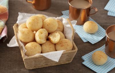 Fotografia em tons de azul em uma bancada de madeira escura, guardanapos de pano azul claro, uma cesta de vime com vários pães de queijo, xícaras cobre com café com leite dentro.
