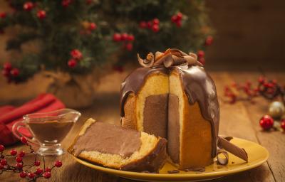 Foto frontal de um panettone com uma fatia cortada. Ele está sobre um prato amarelo e dentro há um recheio de sorvete em tom marrom escuro. Na bancada de madeira há elementos de natal e um pequeno recipiente com calda de chocolate