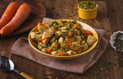 Imagem de uma bancada de madeira escura, sobre a qual há um prato amarelo com a receita de refogado de frango, outro prato com duas cenouras dentro e um pote pequeno com ervilhas verdes ao fundo