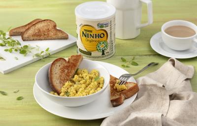 Fotografia em tons de verde e branco de uma bancada verde com um prato branco com um recipiente branco, sobre ele ovos mexidos e pão tostado. Ao lado um paninho beje, uma tábua com pão de forma tostado e orégano, uma lata de NINHO orgânico e uma xícara.