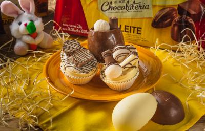 Foto aproximada de três miniovos de chocolate brancos, em um prato amarelo sobre uma bancada de madeira decorada com palha, um tecido amarelo, um pequeno coelho de pelúcia e uma barra de chocolate ao fundo Foto aproximada de três miniovos de chocolate brancos, em um prato amarelo sobre uma bancada de madeira decorada com palha, um tecido amarelo, um pequeno coelho de pelúcia e uma barra de chocolate ao fundo
