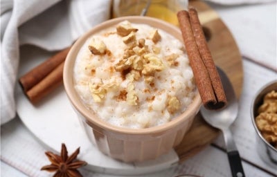 Em cima de uma mesa, recipiente redondo com arroz doce cremoso, com uma textura suave e aveludada, decorado com nozes picadas e canela em pau.