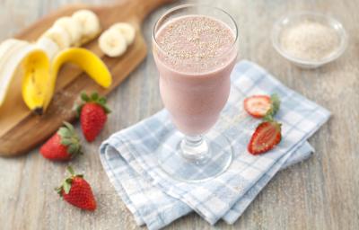 Fotografia em tons de cinza, azul, vermelho e amarelo de uma bancada de madeira cinza com um paninho xadrez azul e branco, sobre ele fatias de morango e um copo de vidro com a vitamina de banana e morango.